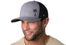 A smiling man with short brown hair and a trimmed beard wears the Haka Hat Palm Tree Trucker Hat, a gray and black Island Snapback with a palm tree logo, paired with a light beige shirt.