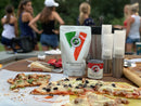 Several homemade pizzas with various toppings sit on a table outdoors, alongside Urban Slicer the WORX! + Sauce from Urban Slicer Pizza Worx. In the background, people are chatting by some greenery.