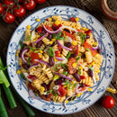 A plate of colorful pasta salad with fusilli, kidney beans, corn, red onion, tomatoes, and green onions on a white and blue plate is finished with SALTOPIA Infused Sea Salts’ Garden Herbs & Lemon Seasoning and surrounded by fresh veggies.