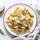 A white plate of creamy penne with mushrooms is topped with SALTOPIA Infused Sea Salts’ GODFATHER Parmesan Black Truffle Seasoning and grated cheese, set on a light surface beside a napkin, fork, and fresh thyme sprigs.