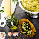 A top-down view of cooked pasta in a colander, a wedge of cheese, a bottle of olive oil, sliced garlic with herbs, garlic cloves, SALTOPIA Infused Sea Salts: MOUTH FULL OF MAGIC garlic salt, and scattered peppercorns on a gray surface.