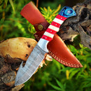 The Texan Knives Custom Handmade Damascus Skinning Knife, featuring an American flag handle, rests on a tree branch beside its brown leather sheath, with green foliage in the background.