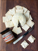A round cheese ball with shredded cheese is surrounded by square crackers on a wooden board, next to a jar of Terrapin Ridge Farms Hot Pepper Peach Jam, a spreading knife, and two crackers topped with cheese.