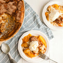 A peach cobbler in a baking dish with two plated servings, each topped with vanilla ice cream and SALTOPIA Infused Sea Salts Caramel Sugar: DATE NIGHT 16oz for extra sweetness. A towel, scoop, and fork sit on a white surface.