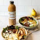 Two bowls of salad with grilled chicken, apples, pecans, and feta rest on a countertop. In the background are a yellow pear and a quart bottle of Terrapin Ridge Farms Apple Cider Maple Balsamic Vinaigrette.