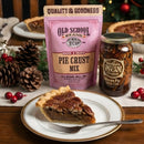 A slice of homemade pecan pie sits before a full pie, the DDR BBQ Supply Classic Southern Pie Pecan Pie Kit, and a jar of filling, surrounded by festive pinecones, berries, and baubles.