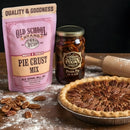 The DDR BBQ Supply Classic Southern Pie Pecan Pie Kit, featuring pie crust mix, pecan filling, loose pecans, a homemade pie, and a rolling pin, is displayed on a floured wooden surface.