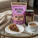 A slice of pecan pie on a white plate, a cup of coffee, the DDR BBQ Supply Classic Southern Pie Pecan Pie Kit bag, and a jar of pecan pie filling rest on a glass table with a cozy knit blanket in the background.
