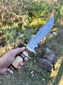 A hand displays the Texan Knives Damascus Steel Sub-Hilt Fighter Bowie Knife 14" outdoors, featuring a patterned blade and polished handle—an ideal Christmas gift that comes with a premium leather sheath.