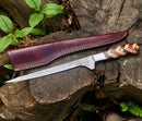 A Handmade Damascus Fillet Knife 8” by Texan Knives, featuring a rosewood and olive wood handle and detailed Damascus blade, rests on a stump beside its leather sheath, surrounded by green plants.