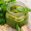 A jar of SALTOPIA Infused Sea Salts PRESTO PESTO Basil, Garlic & Lemon Salt sits amid basil, pine nuts, and garlic cloves on rustic wood, showcasing the fresh flavors inspired by classic pesto sauce.