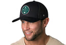 Smiling man with trimmed beard wears a light shirt and a Haka Hat Philadelphia Trucker Hat—Black PHI X Football Snapback featuring a green circular PHI patch and white design, against a white background.