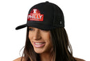 A woman smiles wearing the Haka Hat Philly City of Brotherly Love Trucker Hat, a black cityscape snapback with a red and white PHILLY patch. The background is white.