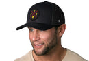 A man with a trimmed beard and mustache smiles and looks to the side against a white background, wearing a Haka Hat Phoenix Trucker Hat (Black PHX X Snapback) with a red and yellow circular logo, paired with a light beige shirt.