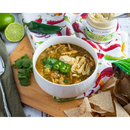 A bowl of chicken chili with jalapeño slices and tortilla strips sits on a wooden board, surrounded by cilantro, lime halves, tortilla chips, and a jar of Terrapin Ridge Farms Hatch Chile Cream Cheese Dip. A colorful napkin is nearby.