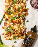 Rectangular flatbread pizza with bacon, chicken, red onion, cheese, and cilantro sits on parchment. Nearby is a bottle of Terrapin Ridge Farms Sweet Heat Grill & Wing Squeeze and fresh green onions with sliced red onions.
