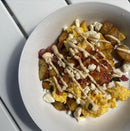 A white bowl on a white table holds scrambled eggs, roasted potatoes, cheese, salsa, and a drizzle of Terrapin Ridge Farms Jalapeno Aioli Squeeze Garnishing Sauce. Sunlight casts shadows across the scene.