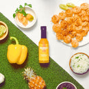 A bottle of Bravado Spice Co Pineapple & Habanero Hot Sauce—a vibrant hot sauce from Bravado Spice Co featuring pineapple and habanero—is surrounded by grilled shrimp, rice, sliced yellow bell pepper, pineapple, pickled ginger, lime wedges, and green garnish on a grass-textured and white background.