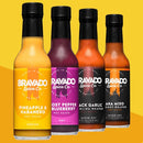 Four bottles of Bravado Spice Co. hot sauce are lined up in a row against a bright yellow background, showcasing bold fruit-infused flavors such as Pineapple & Habanero Hot Sauce, Ghost Pepper Blueberry, Black Garlic Carolina Reaper, and Aka Miso.