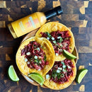 Three tacos filled with shredded meat, onions, and cilantro are served on a plate with lime wedges. Nearby, a bottle of Bravado Spice Co Pineapple & Habanero Hot Sauce from Bravado Spice Co sits on a wooden surface, ready to add its signature sweet and spicy tropical twist to every bite.
