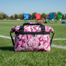The Taiga Pink Camo soft side 18 pack cooler from Taiga Coolers sits on a grassy sports field, with blue, green, and red foldable chairs in the background—perfect for your next outing.