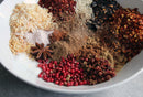 A white bowl showcases World Spice Pink Chili Crisp by World Spice—a vibrant spice blend with colorful piles of dried onion, pink peppercorns, crushed red pepper, star anise, assorted ground spices, sesame seeds, and coarse salt artfully arranged in separate mounds.