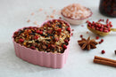 A pink heart-shaped bowl filled with World Spice's Pink Chili Crisp—a vibrant blend of spices and dried herbs—is surrounded by star anise, cinnamon sticks, pink peppercorns, and a bowl of pink Himalayan salt on a light surface.