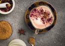 A glass cup of World Spice Pink Chai by World Spice, topped with frothed milk, rose petals, and a star anise, rests on a decorative saucer. Nearby are a bowl of brown sugar, a spoon, a small pitcher, and a mortar filled with rose petals. This exquisite blend from World Spice enchants the senses with its soothing aroma and beautiful presentation.