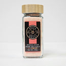 A 145g glass jar of Spice Wheel Himalayan Pink Sea Salt by Spice Wheel, harvested from the ancient Himalayan mountains, featuring a black label with gold and white text, a wooden lid, and filled with distinctly pink, mineral-rich salt.