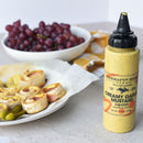 A Terrapin Ridge Farms Creamy Garlic Mustard Squeeze bottle sits beside a plate of ham and cheese pinwheels, pickles, and a bowl of red grapes.
