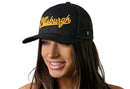 A woman with long brown hair smiles while wearing the Haka Hat Pittsburgh Gold Script Trucker Hat, a black Pennsylvania snapback, featuring "Pittsburgh" in yellow cursive. The background is white.