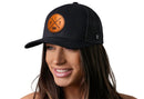 A woman with long brown hair smiles while wearing a Haka Hat Pittsburgh Trucker Hat Leather | Black PIT X Snapback by Haka Hat, featuring a round brown leather patch with ITP letters on the front. The background is white.