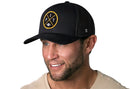 A man with a trimmed beard and blue eyes smiles, wearing a black Haka Hat Pittsburgh Trucker Hat (PIT X Football Snapback) featuring a circular yellow and white emblem, while looking slightly to the side.