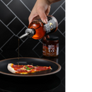 A hand pours Spice Witch Chili Oil from a glass bottle onto pepperoni pizza on a black plate, with a jar of Spice Witch Chili Crisp in the background against a black tiled wall.