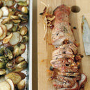 Sliced roasted pork tenderloin on a wooden cutting board, seasoned with SALTOPIA Infused Sea Salts' Saltopia Garlic & Onion Salt: ROOT MATES, beside roasted Brussels sprouts and sliced potatoes, with a knife on the side.