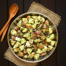 A bowl of potato salad with parsley, red onions, and herbs, seasoned with SALTOPIA Infused Sea Salts’ MUSTARD BEEN LOVE French Mustard Salt, sits on burlap beside wooden salad servers on a dark wooden table.