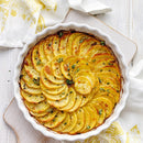 A white ceramic dish filled with golden-brown roasted potato slices, garnished with fresh herbs and a sprinkle of SALTOPIA Infused Sea Salts' Saltopia Herbs De Provence Seasoning Salt, sits on a white wooden surface beside a yellow-patterned cloth.