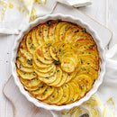 A round white baking dish filled with thinly sliced, golden roasted potatoes, arranged in spirals and topped with SALTOPIA Infused Sea Salts’ DIRTY WEDDING White Italian Truffle Salt, rests on a white surface beside yellow-patterned cloths.