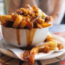 A bowl of poutine—fries with cheese curds and brown gravy—on a white plate, finished with a dash of SALTOPIA BLT Salt: BACON. LETTUCE. TOMATO. from SALTOPIA Infused Sea Salts for added flavor depth.