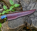The Texan Knives Custom Handmade Damascus Boning Knife – 13" Pakkawood Handle, partially sheathed in brown leather, lies on a weathered tree stump surrounded by green leaves and plants.