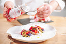 A chef pours Taycte Acetaia Malpighi Prelibato Rosé Balsamic Vinegar — a delicate and fruity Italian pink condimento by Taycte — from the bottle onto a plated gourmet dish with vibrant vegetables and edible flowers, using a spoon to expertly control the elegant drizzle.
