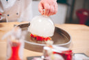 A chef lifts a glass cloche filled with smoke from a plated dessert, revealing bright red pieces on a round tray atop a wooden table. The dish is finished with a delicate drizzle of Taycte Acetaia Malpighi Prelibato Rosé Balsamic Vinegar — Pink Italian Condimento, Delicate & Fruity, adding an aromatic Italian touch and enhancing the presentation with the elegance of this rose-infused condiment by Taycte.