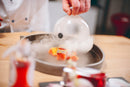 A chef lifts a glass dome, letting smoke swirl around a plate of gourmet cuisine featuring vibrant garnished salmon pieces drizzled with a rose-infused dressing crafted from Taycte Acetaia Malpighi Prelibato Rosé Balsamic Vinegar—a delicate and fruity pink Italian condimento—presented on a wooden table.