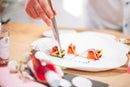 A chef uses tweezers to carefully plate colorful, gourmet food on a white dish, finishing the elegant presentation with a delicate drizzle of Taycte Acetaia Malpighi Prelibato Rosé Balsamic Vinegar—Pink Italian Condimento known for its fruity notes—all arranged beautifully on a wooden surface.