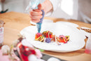 A chef decorates a gourmet dish with a delicate pink sauce—using a piping bag filled with Taycte Acetaia Malpighi Prelibato Rosé Balsamic Vinegar, a fruity Italian condimento—on a white plate surrounded by colorful vegetables on a wooden table.