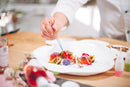 A chef uses tweezers to carefully arrange colorful, gourmet food on a white plate, showcasing an artistic and precise plating technique atop a wooden table. A drizzle of Taycte Acetaia Malpighi Prelibato Rosé Balsamic Vinegar — a pink Italian condimento known for its delicate and fruity flavor — adds an elegant touch and subtle floral note to the presentation.