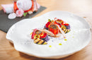 Two pieces of seared fish with grilled zucchini, purple mashed potatoes, flower petals, and delicate sauce drops—highlighted by a rose-infused drizzle of Taycte Acetaia Malpighi Prelibato Rosé Balsamic Vinegar (a pink Italian condimento that’s both delicate and fruity)—are artfully arranged on a white plate. In the softly blurred background, a bottle of this signature Taycte vinegar and fresh flowers add an elegant touch.
