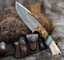 The Texan Knives Premium Damascus Guthook Skinning Knife features a patterned 10” blade and a rosewood & bone handle, resting on bark next to its brown leather sheath—a top hunting essential for any outdoor adventure.