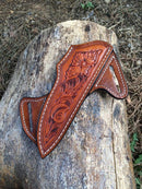 The Texan Knives Premium Bull Leather Knife Sheath for Cowboy Knives, featuring floral patterns and white stitching, rests on a weathered log outdoors among dry leaves—an ideal accessory for your cowboy knives.