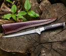 The Texan Knives Premium Custom Handmade Damascus Steel Fillet Knife – 13” Rosewood Handle, with a patterned blade, sits on a tree stump beside its brown leather sheath. Green leaves appear in the background.